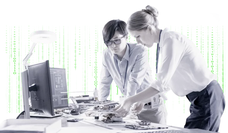 Dwie kobiety pochylone nad biurkiem pełnym urządzeń. Na biurku kilka monitorów. W tle liczby 0 i 1 lecące rzędami w dół