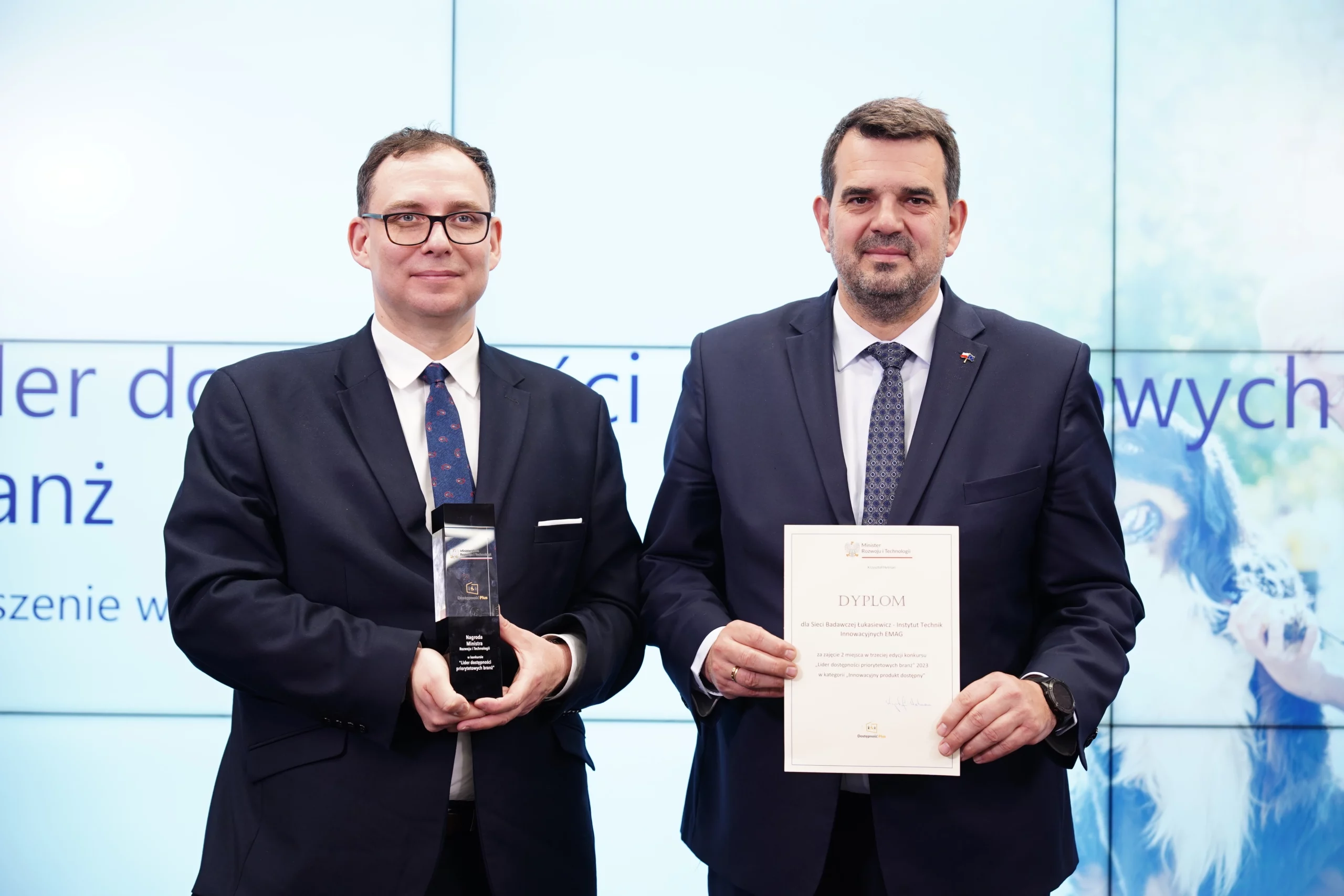 Dwaj mężczyźni w garniturach. Jeden trzyma statuetkę, drugi dyplom