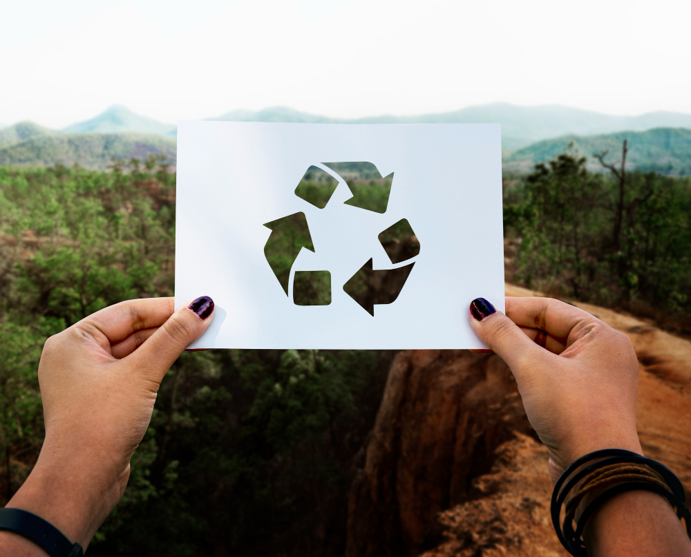 wycięty w kartce symbol recyklingu obrazujący trzy strzałki w kręgu. Kartka trzymana w dłoniach na tle krajobrazu