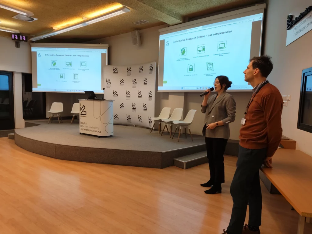 Dwie osoby obok sceny - mężczyzna i kobieta. Kobieta przemawia do mikrofonu.