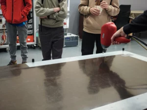 Grupa osób wokół stołu. Przy stole pokaz urządzenia trzymanego w ręku.