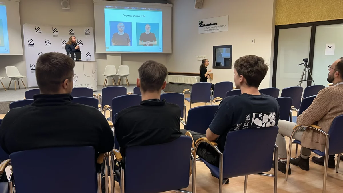 Sala konferencyjna. Na sali prowadzący i zebrani słuchacze. Dodatkowo przy scenie kobieta tłumacząca na Polski Język Migowy.