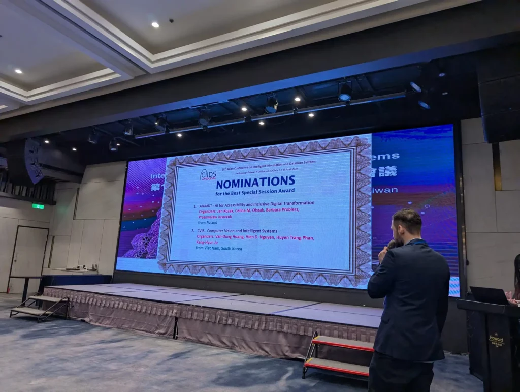 Duża sala z ekranem projektora. Na ekranie napis: Nominacje. Przed ekranem mężczyzna z mikrofonem.